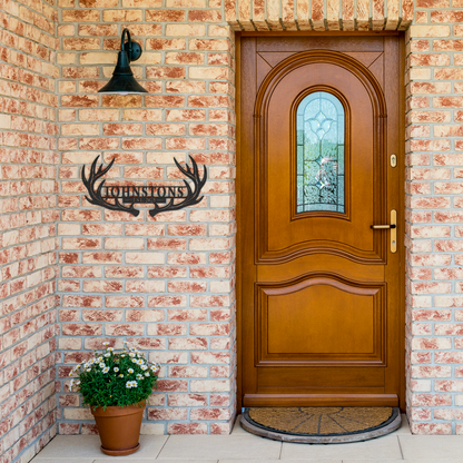 Black custom deer antler family name metal sign by brick entrance — personalized rustic cabin wall decor